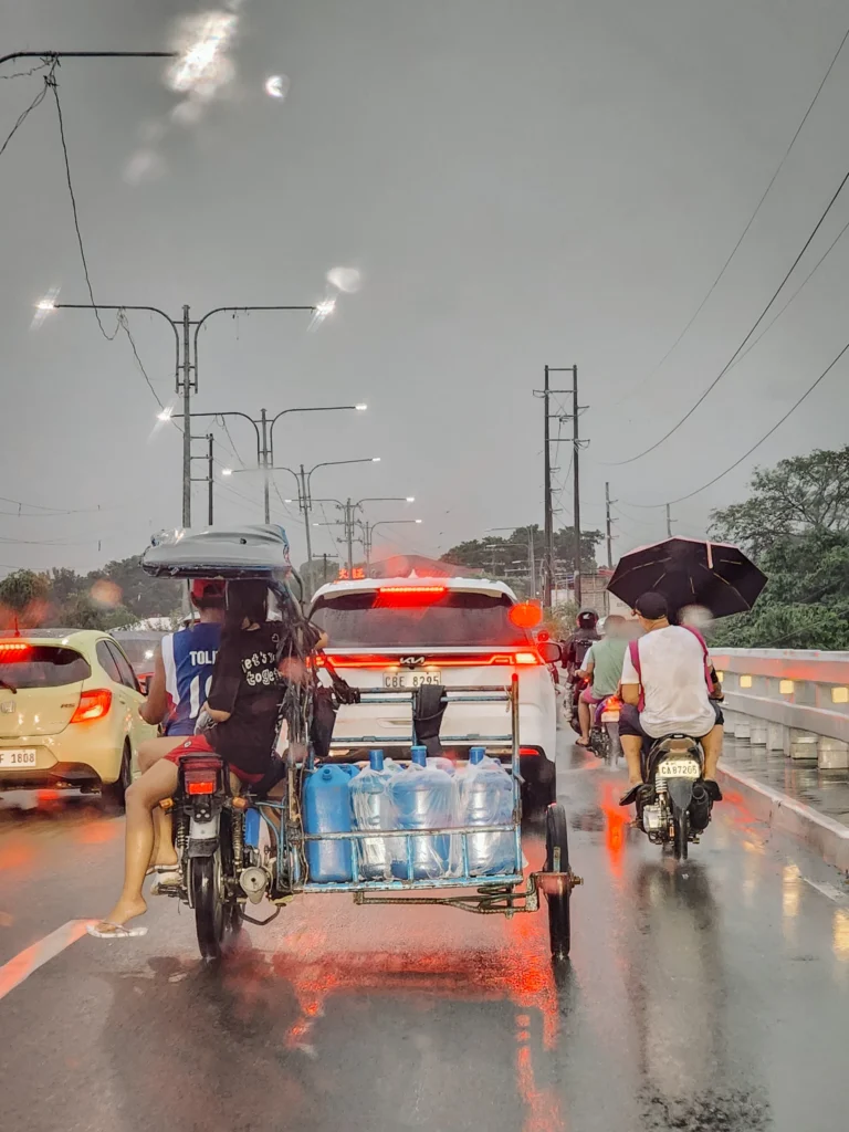 A busy street in Angeles City, Philippines, during a rainy day. A tricycle with several passengers and large water jugs in the sidecar is visible, with a white SUV in front and motorbikes with passengers holding umbrellas in the background. The road is wet, reflecting the city lights and vehicles.