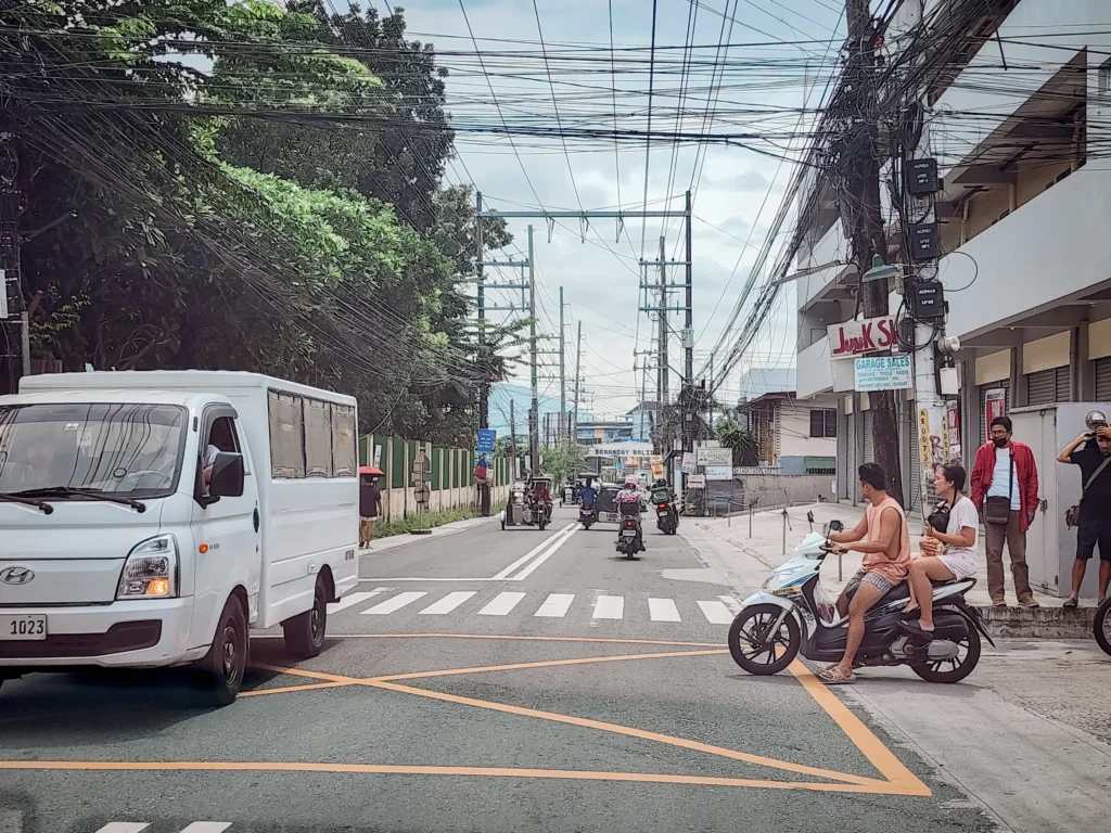 A busy street scene in Angeles City, Philippines, showing a variety of vehicles including a white Hyundai van and motorbikes waiting at a crosswalk. The street is lined with utility poles tangled with numerous electrical wires and trees. People are visible on the sidewalk and a mountainous backdrop is in the distance.
