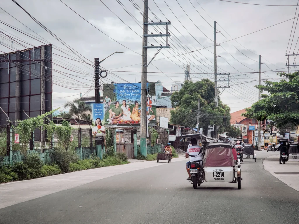 A wide-angle shot of a street in Angeles City, Philippines, showing several tricycles driving away from the camera. The road is lined with utility poles, tangled electrical wires, and large billboards advertising local products. A lush, green landscape and various buildings are visible in the background under a cloudy sky.