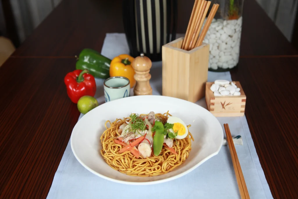A large serving plate filled with Filipino Pancit Canton, showcasing yellow egg noodles mixed with carrots, snap peas, cabbage, shrimp, and sliced meat.