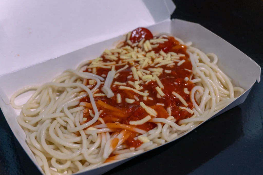 A close-up of a plate of Filipino spaghetti, with bright red sauce, red hot dog slices, and melted orange cheese on top.