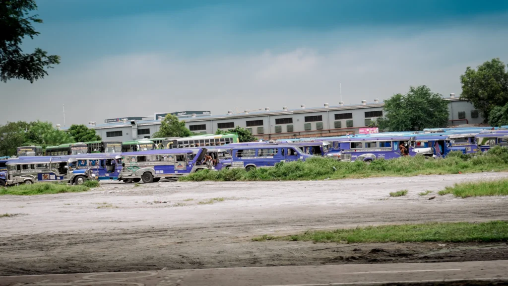 A wide shot of a jeepney terminal in the Philippines, with multiple purple jeepneys parked in rows on a dirt and gravel lot. A large building with a gray facade and a few green trees are visible in the background under a partly cloudy sky.