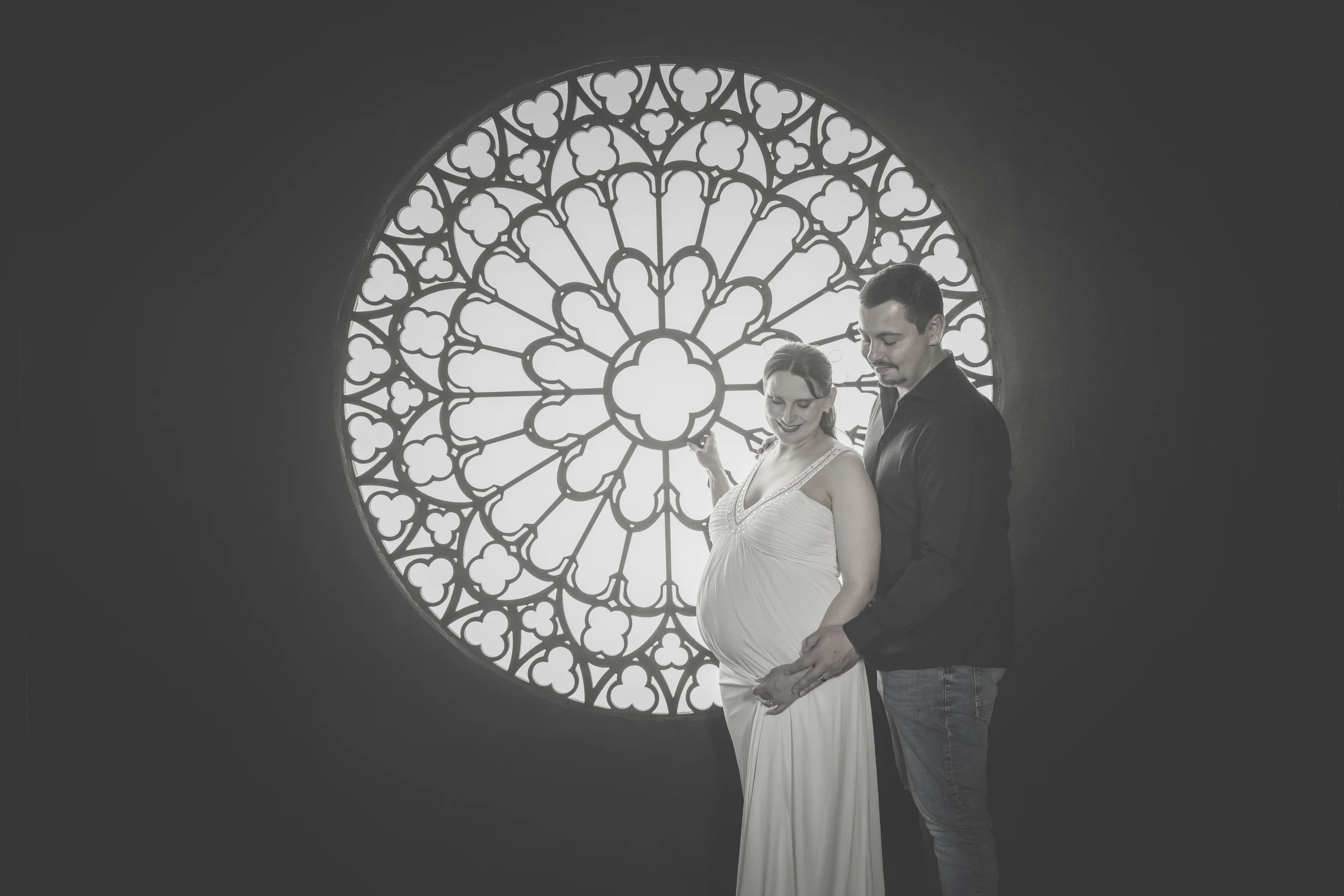 Black and white photo of a pregnant woman and her husband posing in front of a stained-glass window.