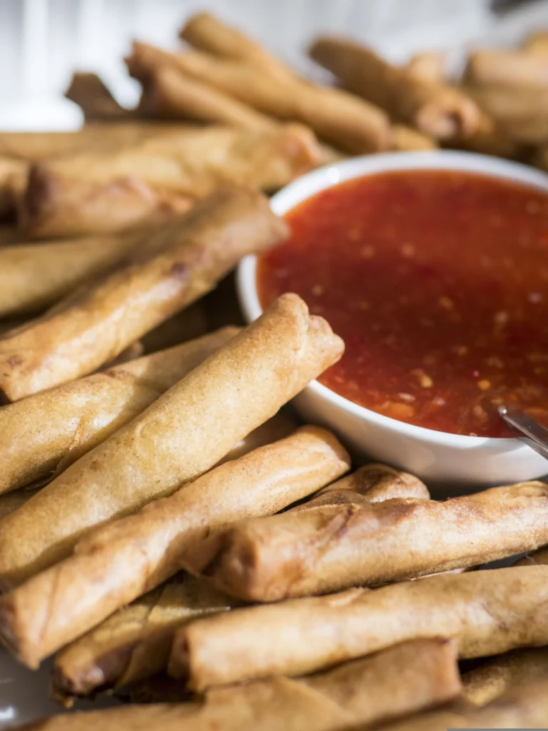 A serving of deep-fried Filipino lumpia (spring rolls) arranged neatly on a plate next to a small bowl of dipping sauce