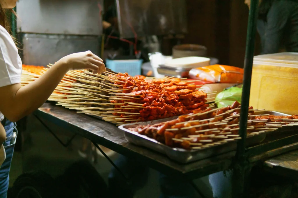 A Filipino street food vendor stands behind their cart, which is stocked with an array of popular street food items like skewers and fried snacks.