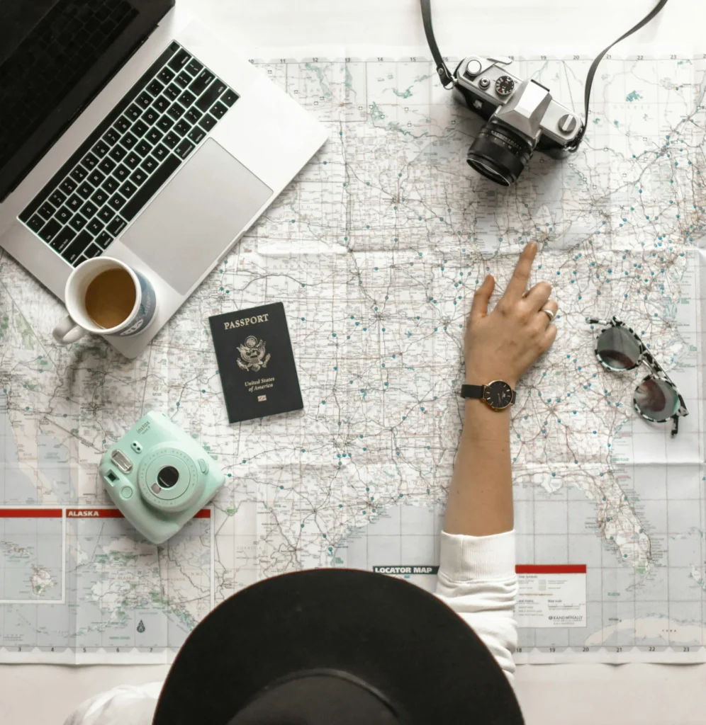 Overhead shot of a person pointing at a map with a passport, laptop, camera, and coffee nearby, symbolizing the planning stage of moving abroad.