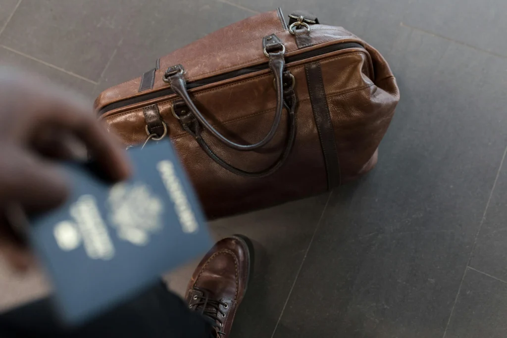 A close-up shot of a hand holding a blue passport, with a blurred brown leather duffel bag on the floor, symbolizing the essential documents needed for moving abroad.