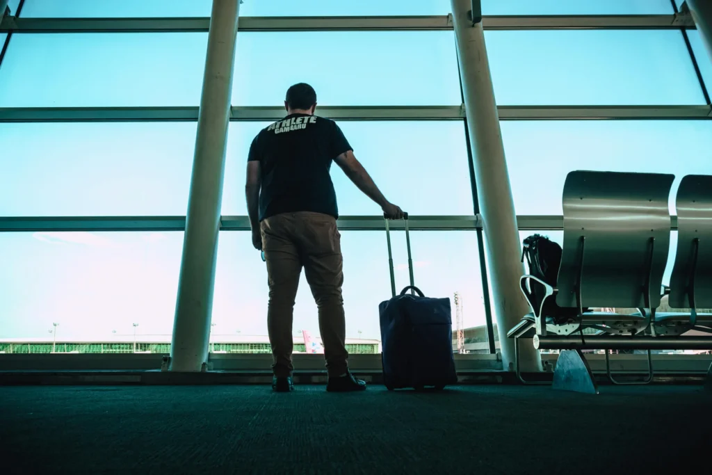 A man seen from behind, standing with his hand on a rolling suitcase, looking out a large airport window at the sky and terminal, symbolizing the journey of moving abroad.