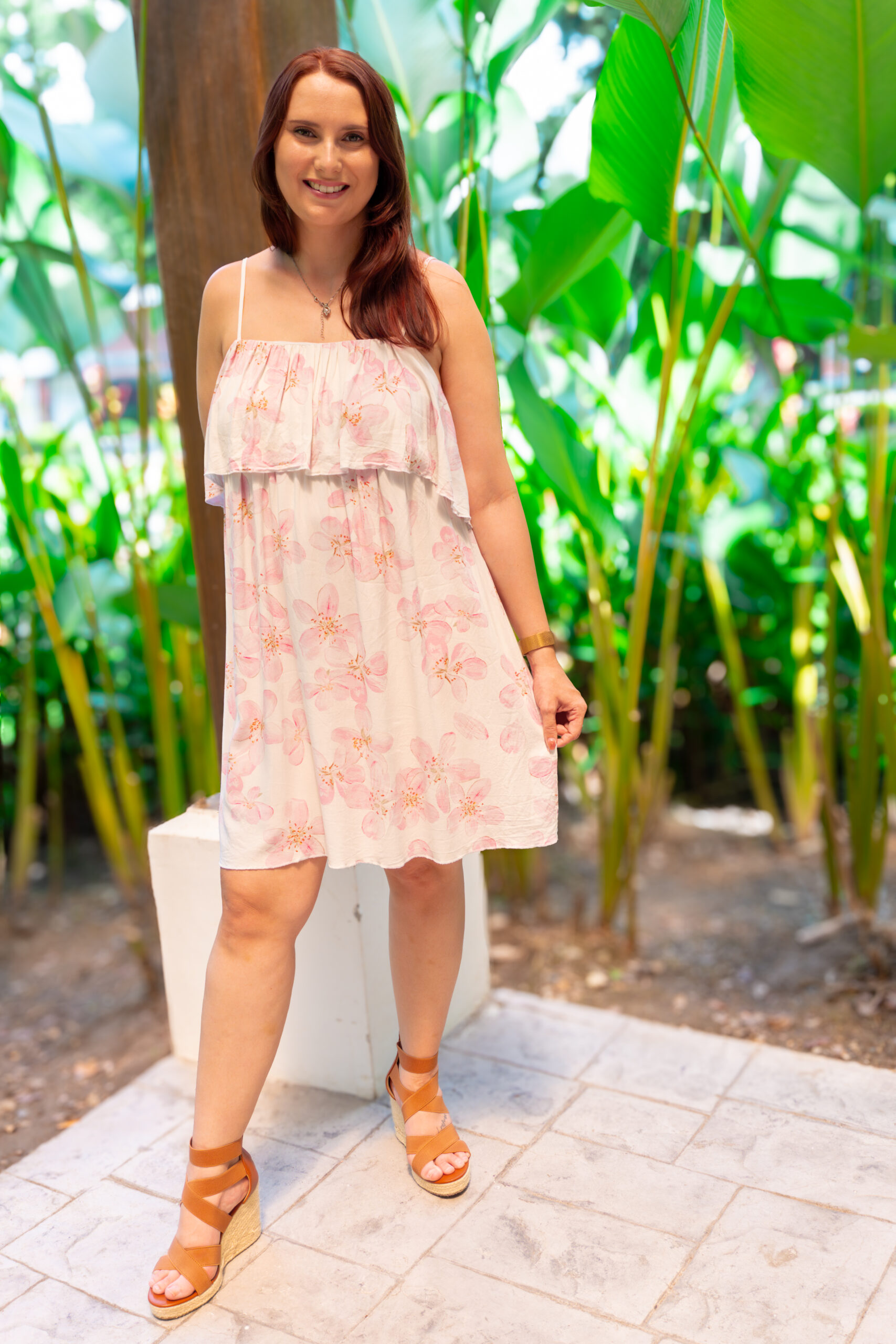 Woman in a light pink floral sundress standing outdoors surrounded by lush green plants, smiling at the camera.